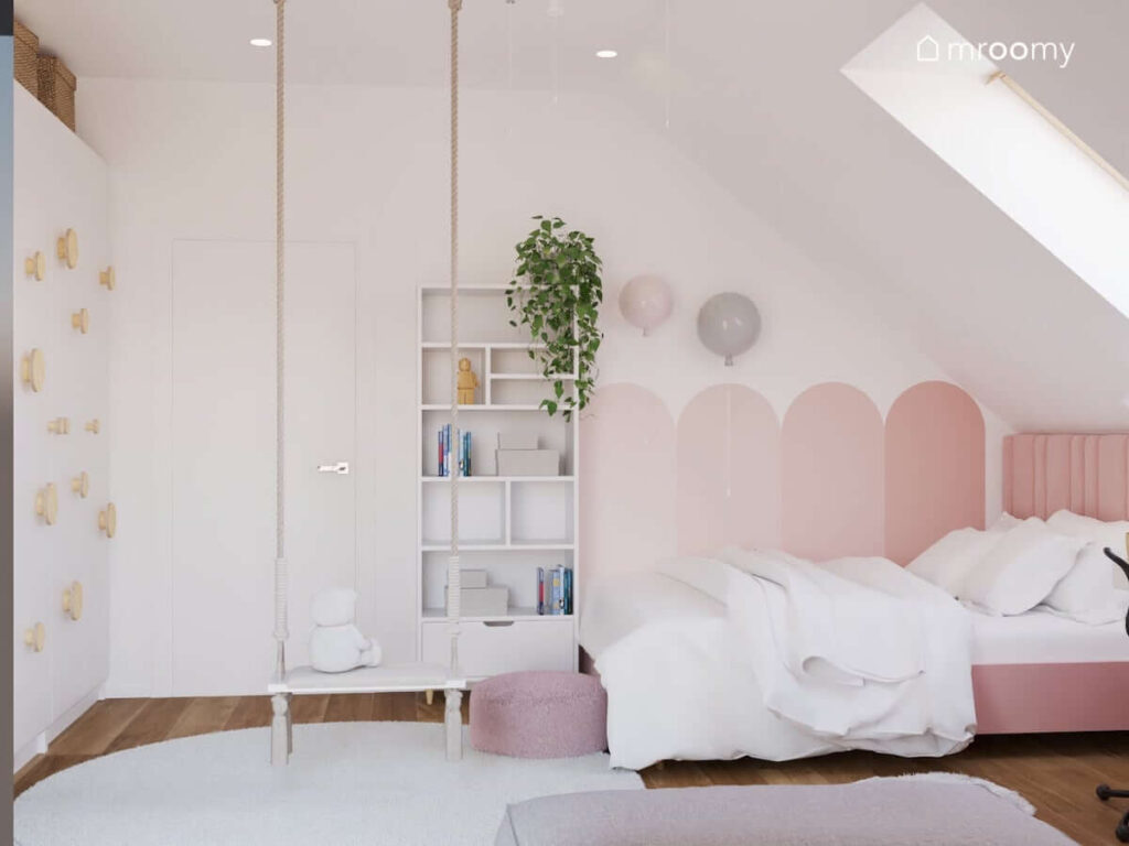 Biały pokój dla dziewczynki z wyrazistymi różowymi dodatkami w postaci łóżka i fragmentu ściany oraz z huśtawką wiszącą i szafą uzupełnioną uchwytami ze sklejki