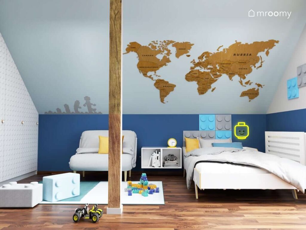 Biało niebieski poddaszowy pokój dla chłopca z białym łóżkiem szarą sofą pufkami oraz panelami ściennymi w kształcie klocków a także drewnianą mapą świata i zabawną naklejką ścienną z Lego i ledonem w kształcie głowy ludzika