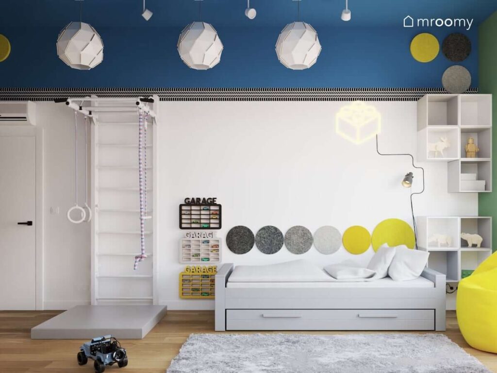Biało niebieski pokój dla chłopca z łóżkiem z szufladą drabinką gimnastyczną i materacem oraz piętrowymi półkami na samochody a także z białymi lampami sufitowymi w geometrycznym kształcie
