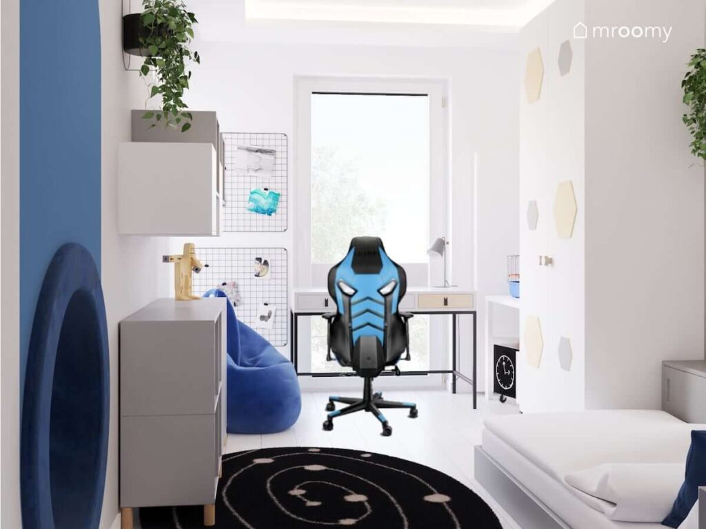 Strefa nauki w pokoju dla chłopca z biurkiem na czarnym stelażu dwoma organizerami ściennymi oraz fotel gamingowym a także niebieska pufa sako i czarno biały dywan