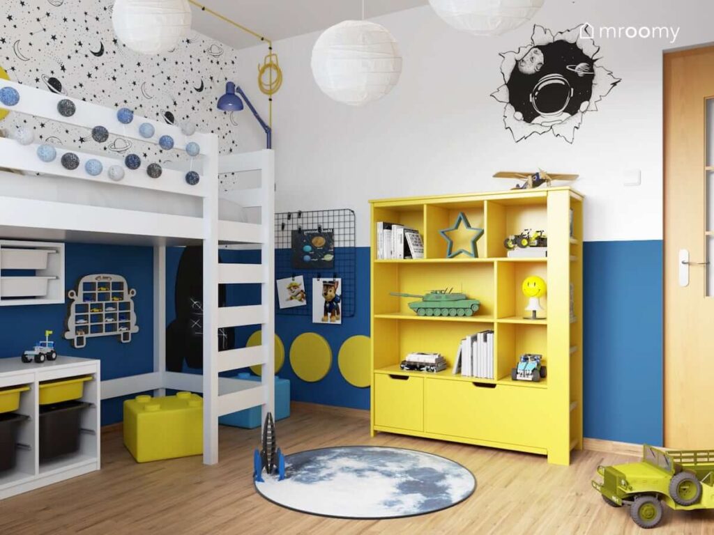 Biało niebieski kosmiczny pokój dla przedszkolaka z białymi i żółtymi meblami naklejką ścienną z kosmonautą kosmiczną tapetą a także niebieską girlandą cotton balls i półką na samochody w kształcie samochodu