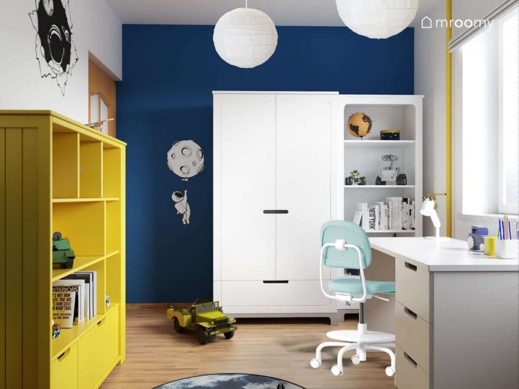Biało niebieski pokój dla chłopca z białą szafą i żółtym regałem a także białym biurkiem i biało niebieskim krzesłem i naklejką z kosmonautą