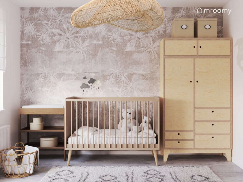 Drewniane łóżeczko z mobilem z chmurkami szafa ze sklejki a na niej kartonowe pojemniki a także drewniany przewijak i tapeta w palmy w pokoju dla noworodka