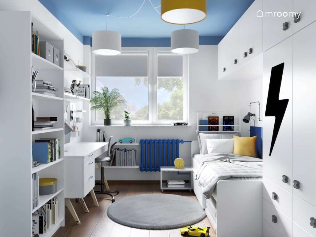 Biało niebieski pokój dla chłopca z białym regałem biurkiem łóżkiem i szafą modułową oraz puszystym szarym dywanem na podłodze i lampą sufitową z abażurami w różnych kolorach