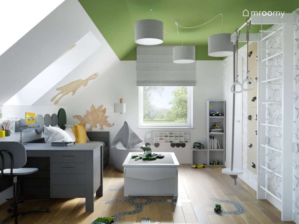 Biało zielony pokój dla chłopca na poddaszu z szarym biurkiem i łóżkiem a także pufą sako i stolikiem do zabawy oraz ozdobnymi dinozaurami ze sklejki na ścianach