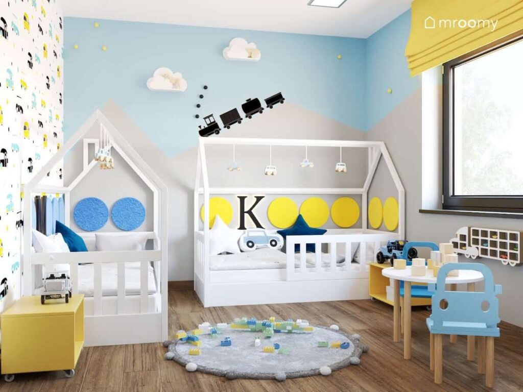 Radosny pokój dla dwóch chłopców z łóżkami w kształcie domków uzupełnionymi żółtymi i niebieskimi panelami ściennymi a także lampkami nocnymi w kształcie literek