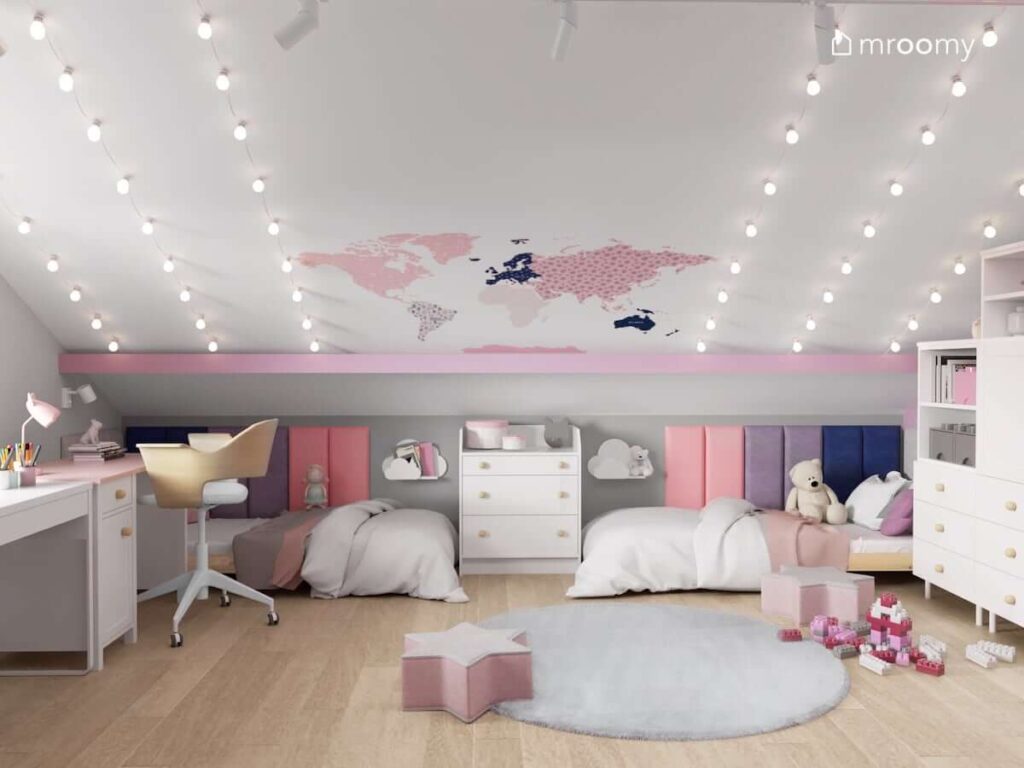 Strefa odpoczynku w pokoju dla dwóch sióstr z niskimi drewnianymi łóżkami uzupełnionymi kolorowymi panelami ściennymi a także skosem ozdobionym naklejką w kształcie mapy świata i sznurami z białych żarówek