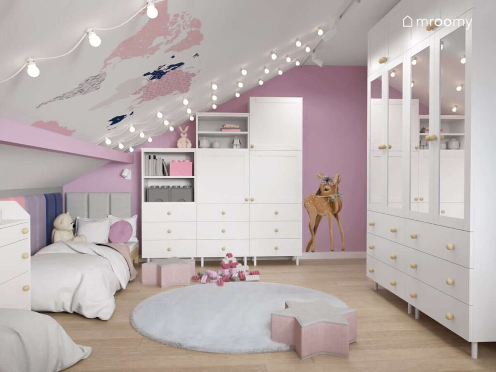 Biało różowy poddaszowy pokój dla dwóch dziewczynek z białymi meblami modułowymi puszystym szarym dywanem i szaro różowymi pufkami w kształcie gwiazdek oraz naklejką ścienną w kształcie sarenki