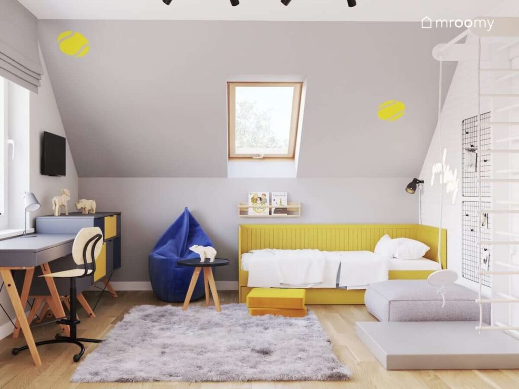 Jasny poddaszowy pokój dla dziewczynki z żółtym łóżkiem szarym biurkiem i komodą niebieską pufą sako oraz skosem ozdobionym naklejkami w kształcie piłek do tenisa