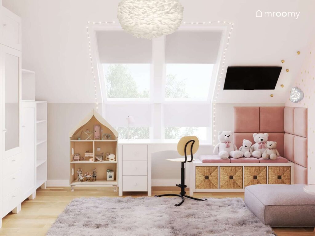 Jasny poddaszowy pokój dla dziewczynki z drewnianą szafką w kształcie domku białym biurkiem pod oknami i niskim regałem uzupełnionym koszami z trawy morskiej oraz z oknami ozdobionymi świetlnymi łańcuchami dekoracyjnymi