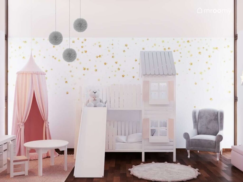 Łóżko z antresolą w kształcie domku baldachim z gniazdkiem do zabawy a także szary fotel i ozdobne pompony oraz tapeta w złote gwiazdki w pokoju dla dziewczynki niemowlaka