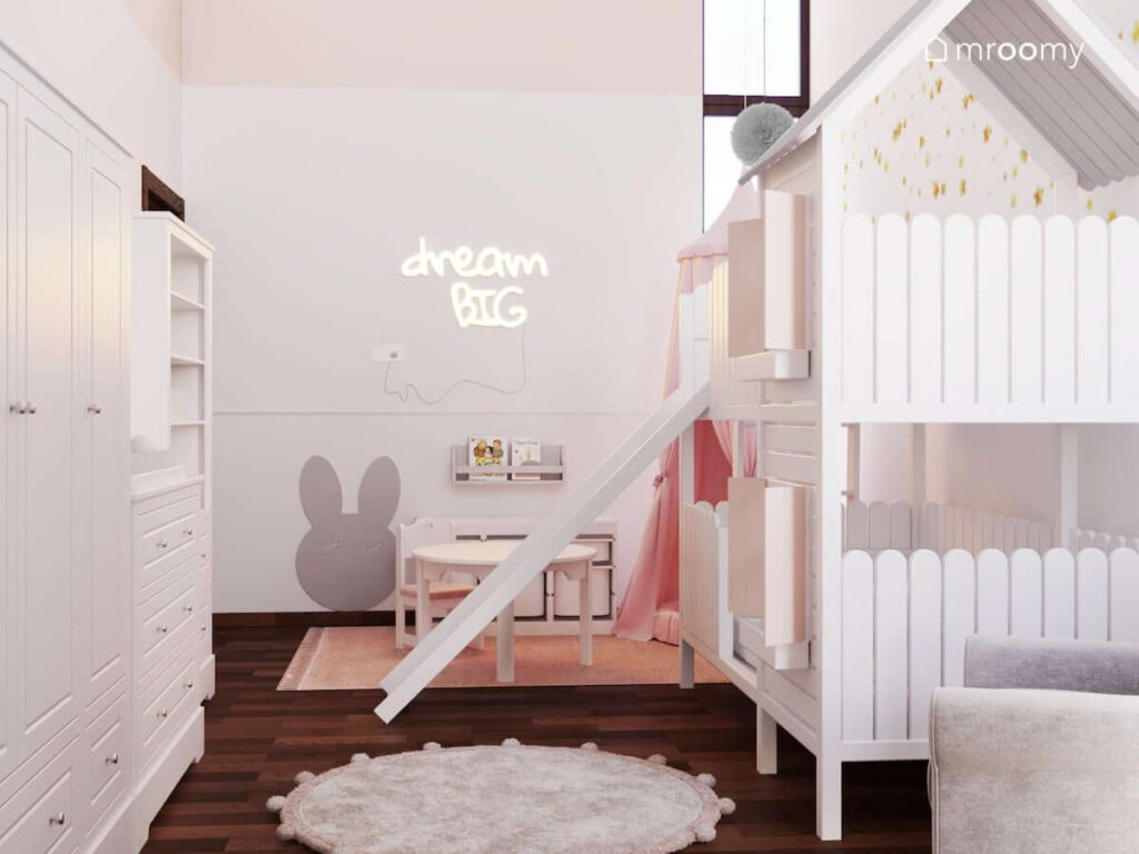 Jasny pokój dla malutkiej dziewczynki z białymi meblami łóżkiem w kształcie domku z także różowym i szarym dywanem i okrągłym drewnianym stolikiem