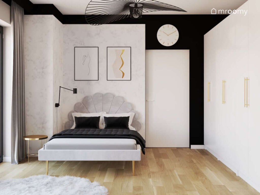 Tapicerowane łóżko z dekoracyjnym zagłówkiem a także ozdobne plakaty i biało złoty zegar w monochromatycznym pokoju dla nastolatki