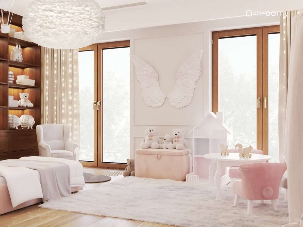 Jasnoróżowy pokój dla dziewczynki z regałem w kształcie domu dla lalek kufrem na zabawki dużym puszystym dywanem a także ozdobnymi skrzydłami na ścianie i dużą dekoracyjną lampą sufitową z piór