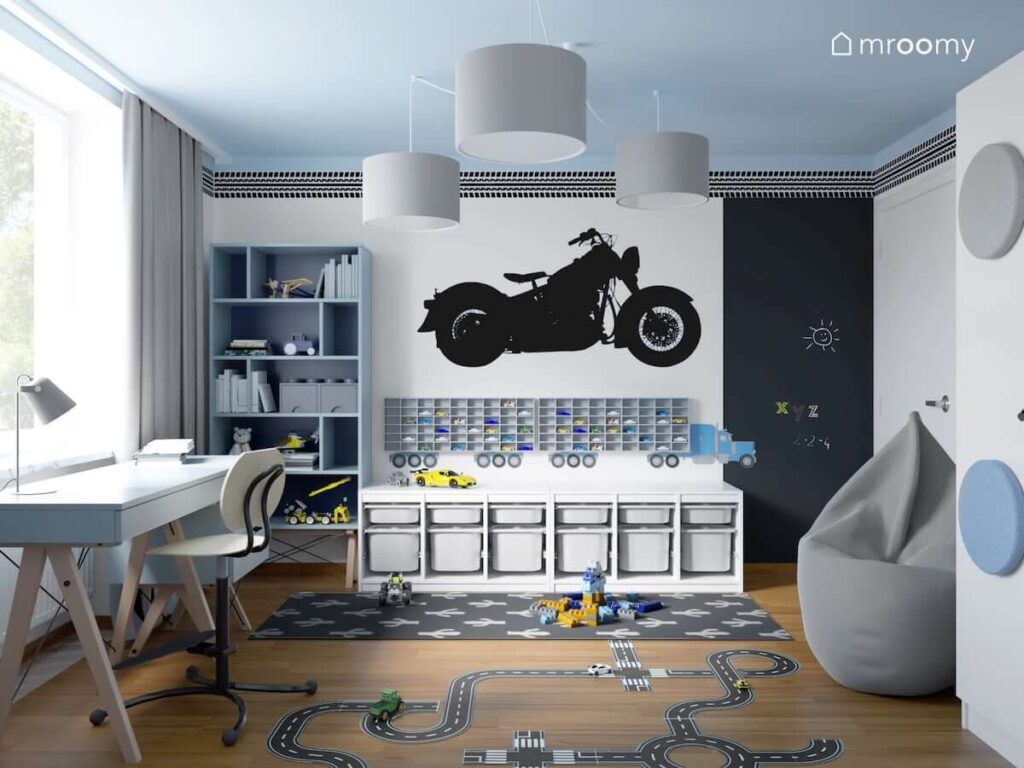 Biało niebieski pokój dla fana motoryzacji z dużą naklejką ścienną w kształcie motocykla oraz naklejką w kształcie śladu opon a także półką na samochody w kształcie tira oraz białym regałem z pojemnikami na zabawki
