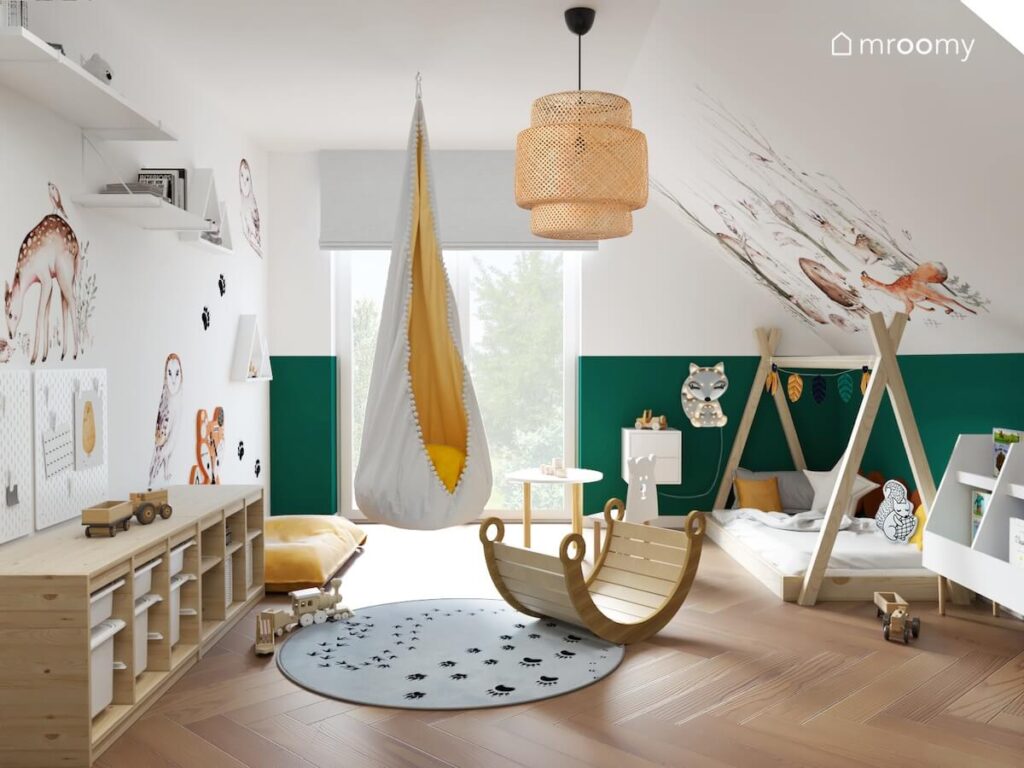 Biało zielony pokój dla chłopca z licznymi motywami zwierzęcymi drewnianym łóżkiem tipi drewnianym regałem z pojemnikami na zabawki a także bambusową lampą i wiszącym fotelem kokonem