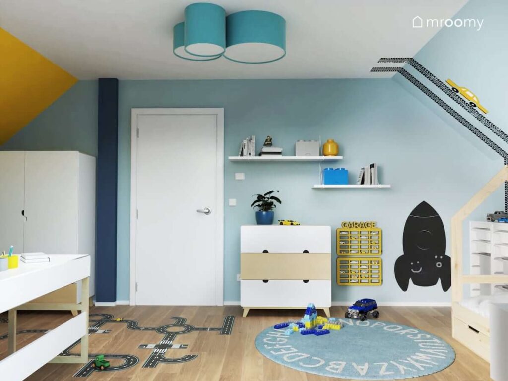 Biało błękitny pokój dla chłopca w wieku przedszkolnym z żółtymi półkami na samochody tablicą kredową w kształcie rakiety oraz niebieską plafoniadą na suficie