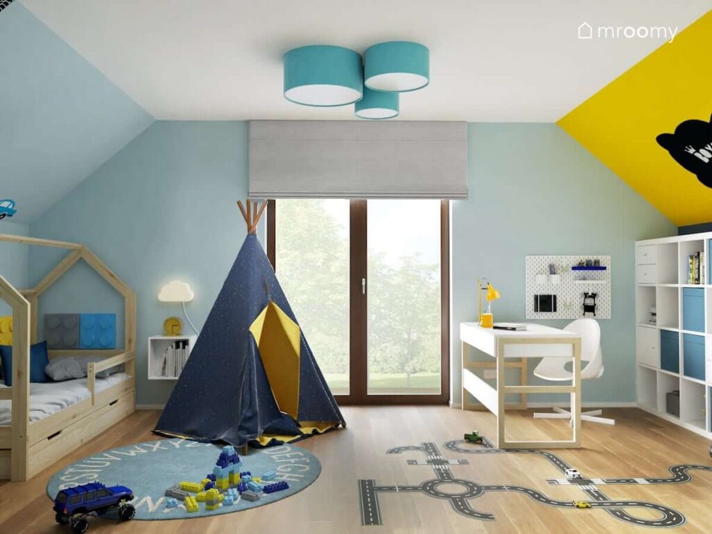 Granatowo musztardowy namiot tipi a także drewniane łóżko domek biało drewniane biurko a na podłodze dywan z alfabetem i naklejka w kształcie jezdni w pokoju dla chłopca