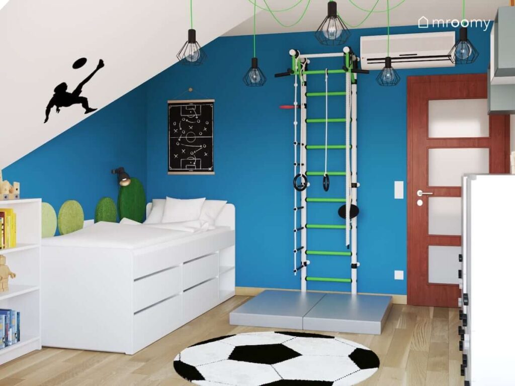 Biało niebieski pokój dla chłopca z drabinką gimnastyczną białym łóżkiem z szufladami a także dywanem w kształcie piłki do futbolu oraz lampą sufitową na ozdobnym zielonym zawieszeniu