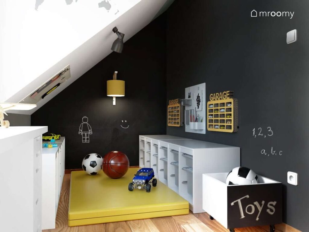 Kącik zabawy w pokoju dla dwóch braci ze ścianami pomalowanymi czarną farbą kredową oraz z półkami na samochody regałami z pojemnikami na zabawki i materacem gimnastycznym