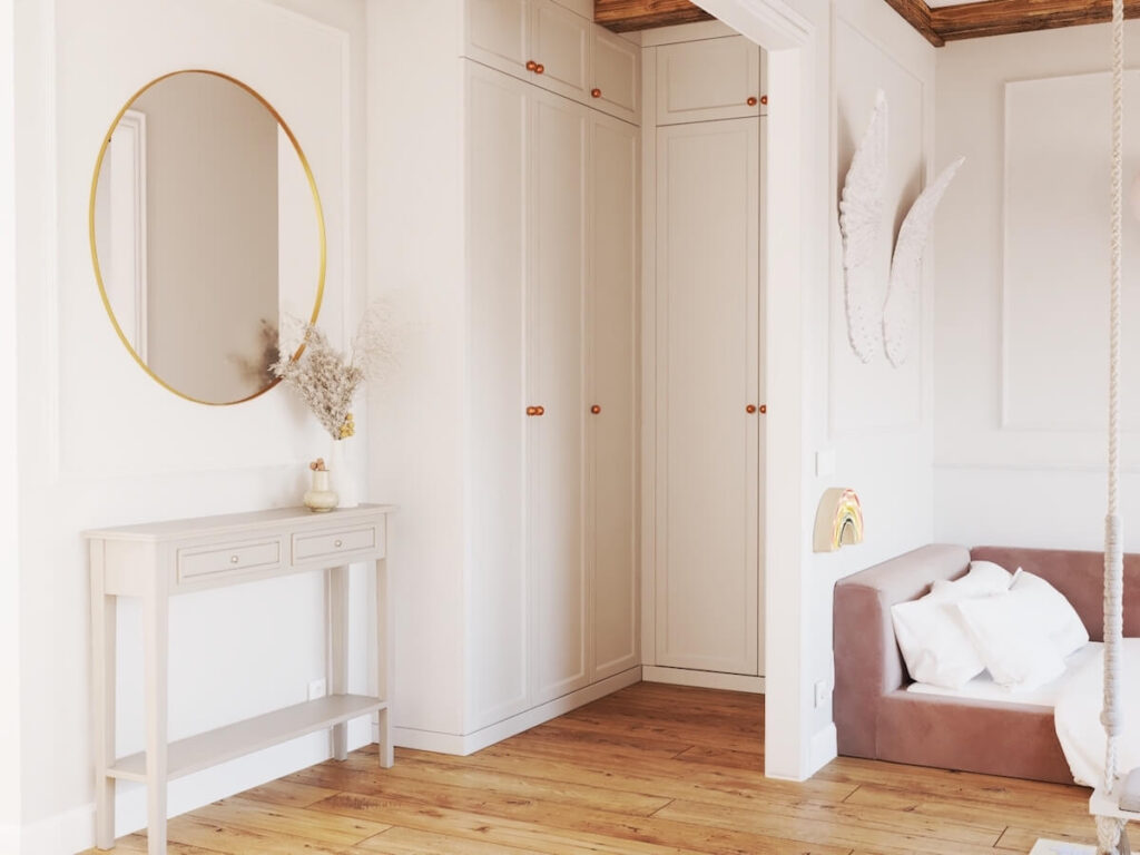 Duże białe szafy a także szara konsola a nad nią lustro w mosiężnej ramie w pokoju dla dziewczynki