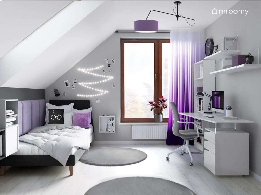 Biało szary pokój dla nastolatki z czarnym tapicerowanym łóżkiem białym biurkiem ozdobnymi lampkami LED oraz z fioletowymi akcentami w postaci poduszki lampy sufitowej i zasłon