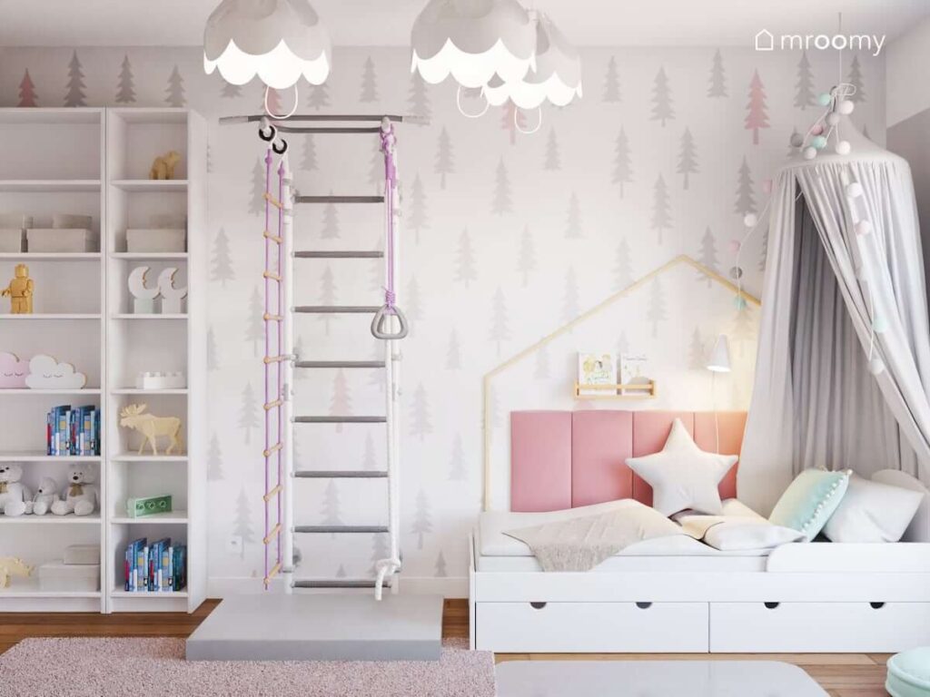 Białe łóżko uzupełnione szarym baldachimem oraz różowymi panelami ściennymi a także wysokie regały i drabinka gimnastyczna a na ścianie delikatna tapeta w choinki w pokoju dla dziewczynki