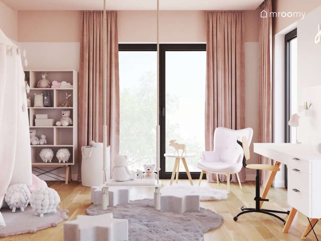 Pudrowo różowy pokój dla dziewczynki z białymi i różowymi meblami na drewnianych nogach oraz wiszącą huśtawką oraz z puszystymi dywanami na podłodze