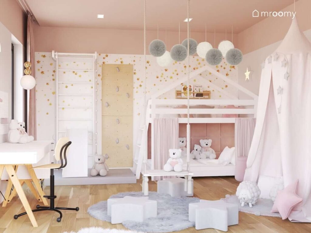 Białe łóżko piętrowe w kształcie domku uzupełnione panelami ściennymi oraz zasłonkami a nad nim białe i szare pompony oraz drabina gimnastyczna i ścianka wspinaczkowa ze sklejki w pokoju dla dziewczynki przedszkolaka