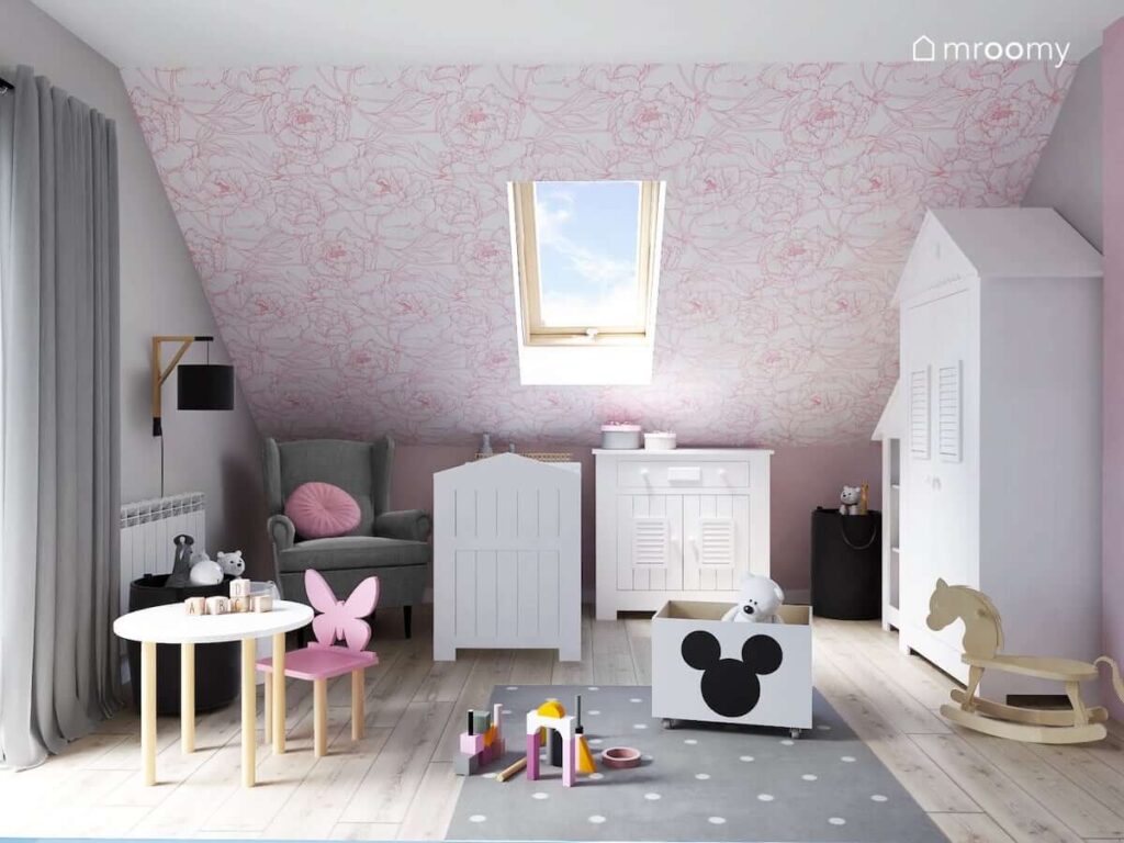 Poddaszowy pokój dla niemowlaka z białymi meblami szarym fotelem i dywanem oraz skosem ozdobionym biało różową tapetą w kwiaty