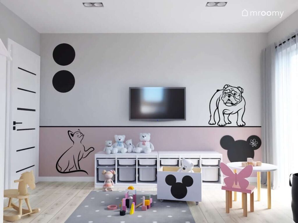 Biało różowy pokój dla malutkiej dziewczynki a w nim regał z pojemnikami na zabawki a także ściana z telewizorem ozdobiona naklejkami w kształcie kota i buldoga