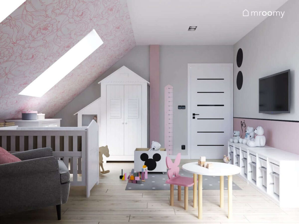 Biało różowy poddaszowy pokój dla niemowlaka z białą szafą w kształcie domku łóżeczkiem a także stolikiem dziecięcym miarką wzrostu i pojemnikami na zabawki