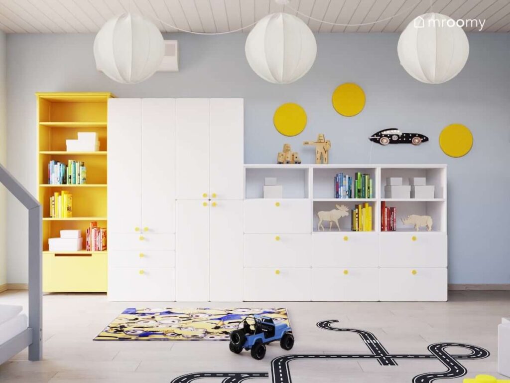 Duża biała szafa modułowa z żółtymi gałkami a także żółty regał a na podłodze dywan z Minionkami i naklejka podłogowa w kształcie jezdni w pokoju dla chłopca