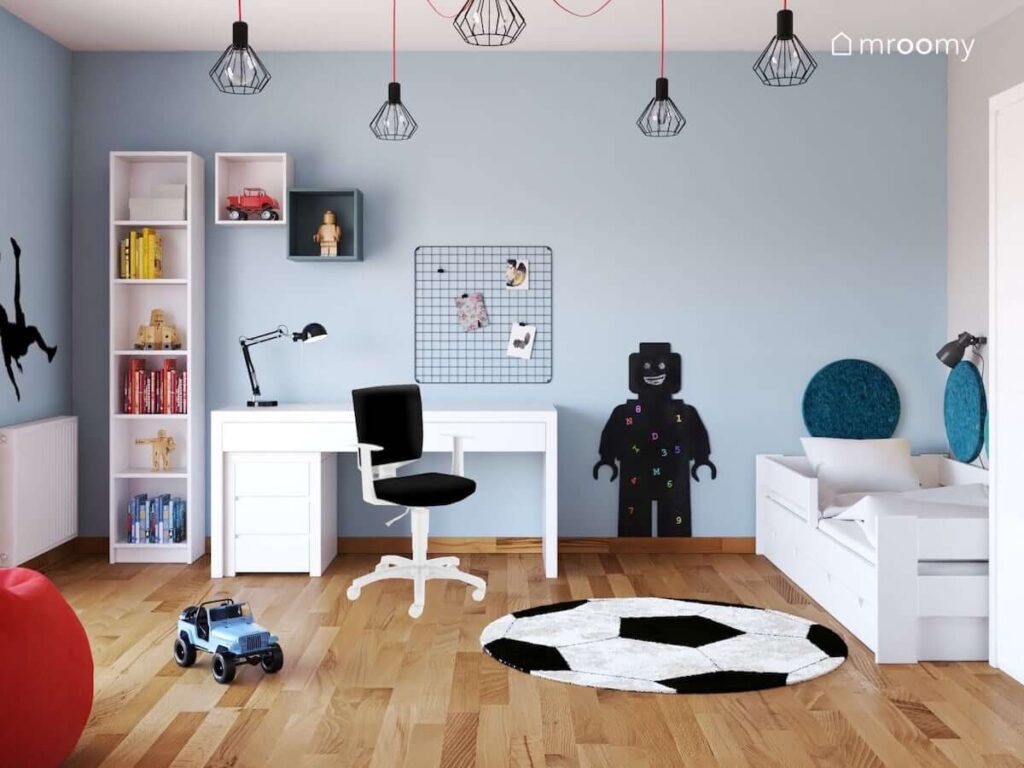 Niebiesko biały pokój dla chłopca z białym biurkiem i regałem a także czarnym organizerem ściennym tablicą kredową w kształcie ludzika lego i dywanem w kształcie piłki