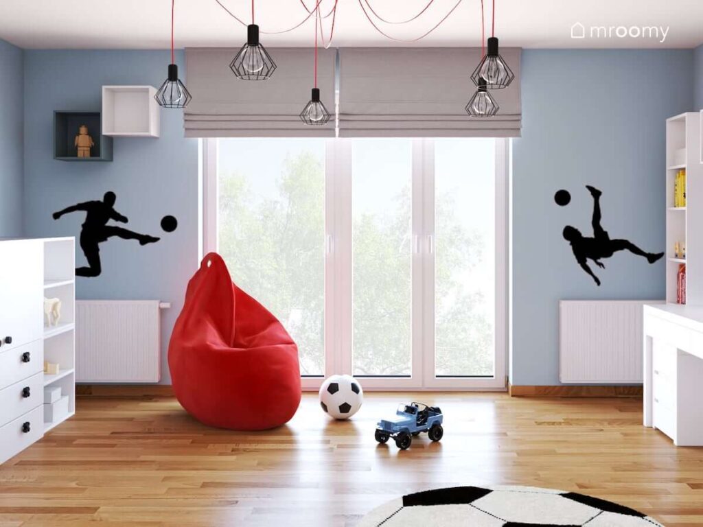 Czerwona pufa sako czerwono czarna lampa sufitowa z rozłożystym zawieszeniem oraz naklejki w kształcie piłkarzy w niebiesko białym pokoju dla fana piłki nożnej