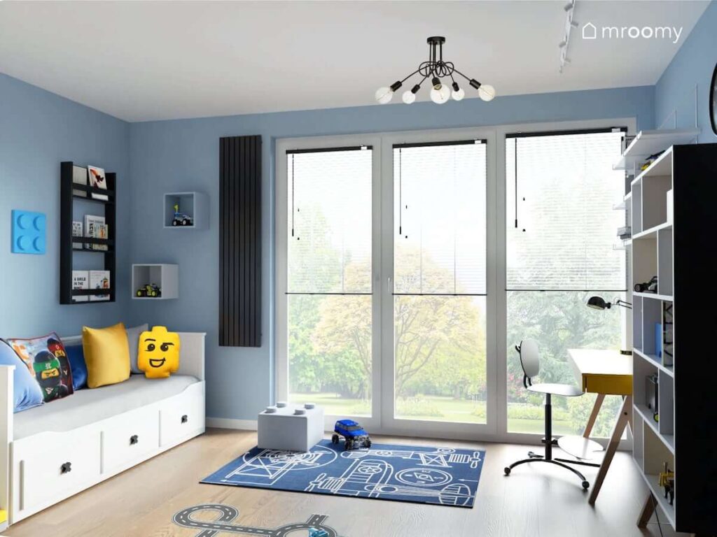 Pokój dla chłopca z niebieskimi ścianami białym łóżkiem z szufladami a także czarną biblioteczką i poduszkami z motywami Lego