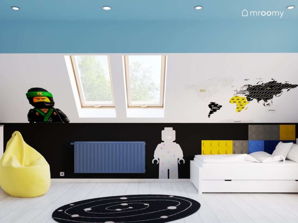 Biała leżanka uzupełniona kolorowymi panelami ściennymi w kształcie klocków a na ścianie naklejka z motywem Lego tablica kredowa w kształcie ludzika Lego i mapa świata