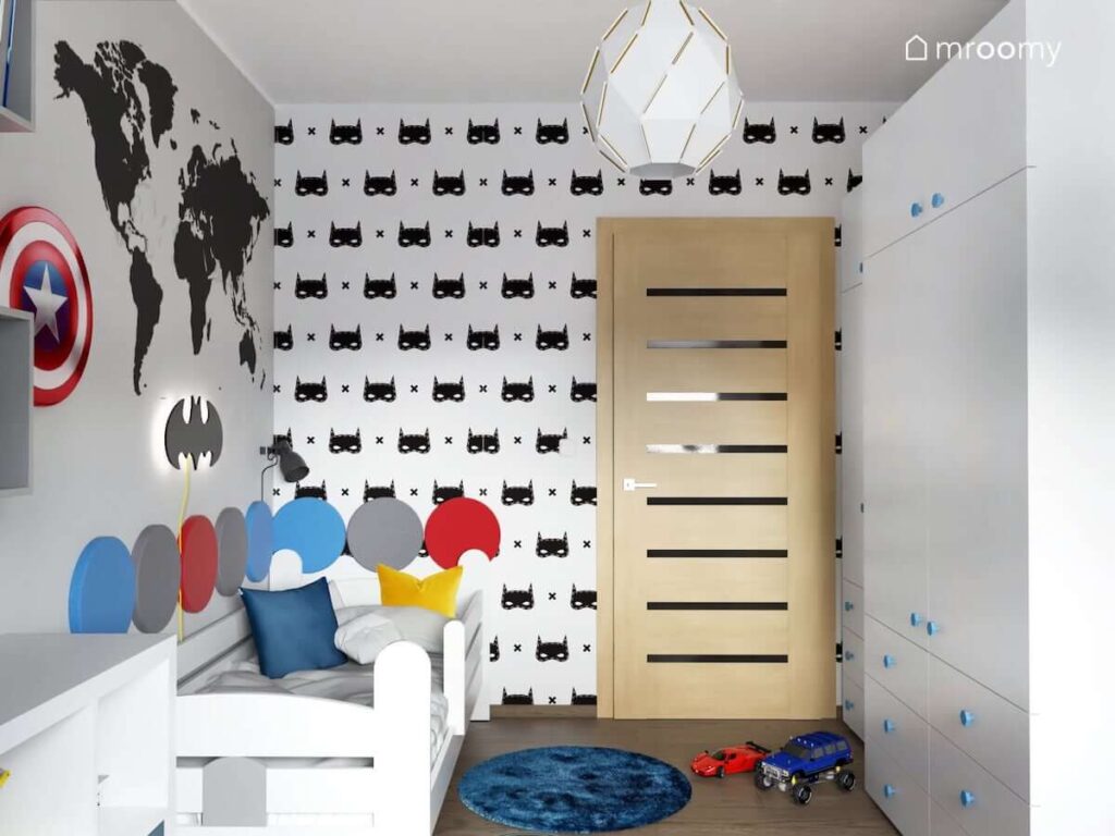 Ściana pokryta tapetą z Batmanem a także białe łóżko uzupełnione panelami ściennymi w różnych kolorach a nad nim mapa świata i naklejka w kształcie logo Batmana