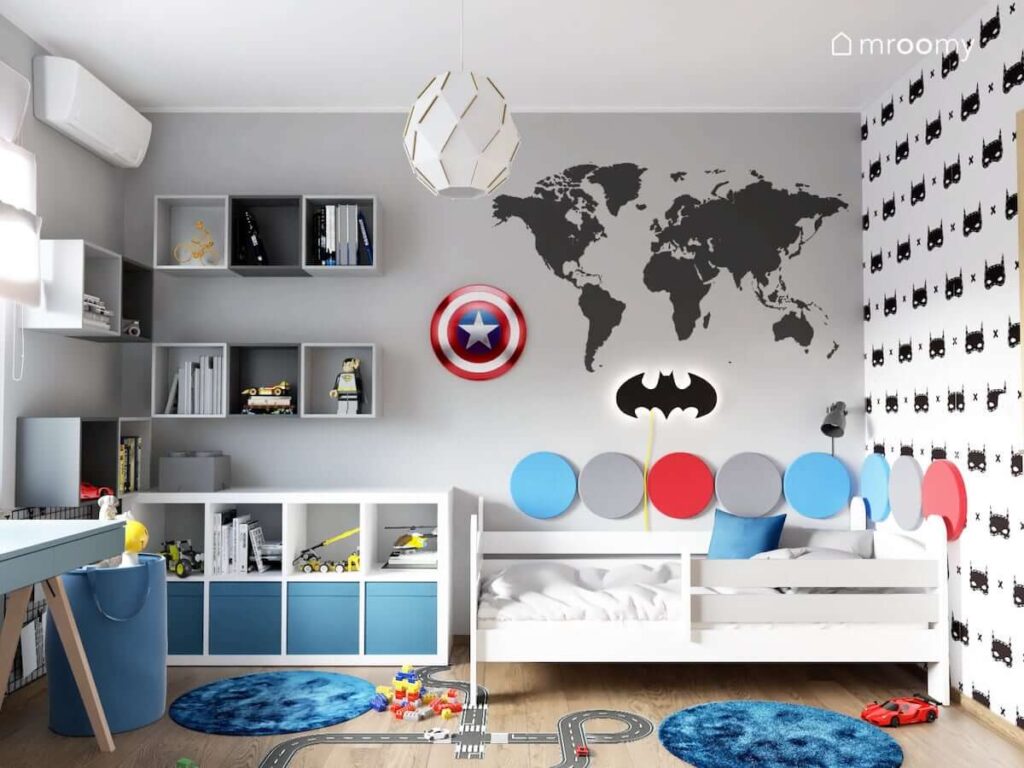 Pokój miłośnika Batmana i Kapitana Ameryki z białym łóżkiem uzupełnionym panelami ściennymi oraz z niskim białym regałem szarymi szafkami ściennymi i niebieskimi dywanami