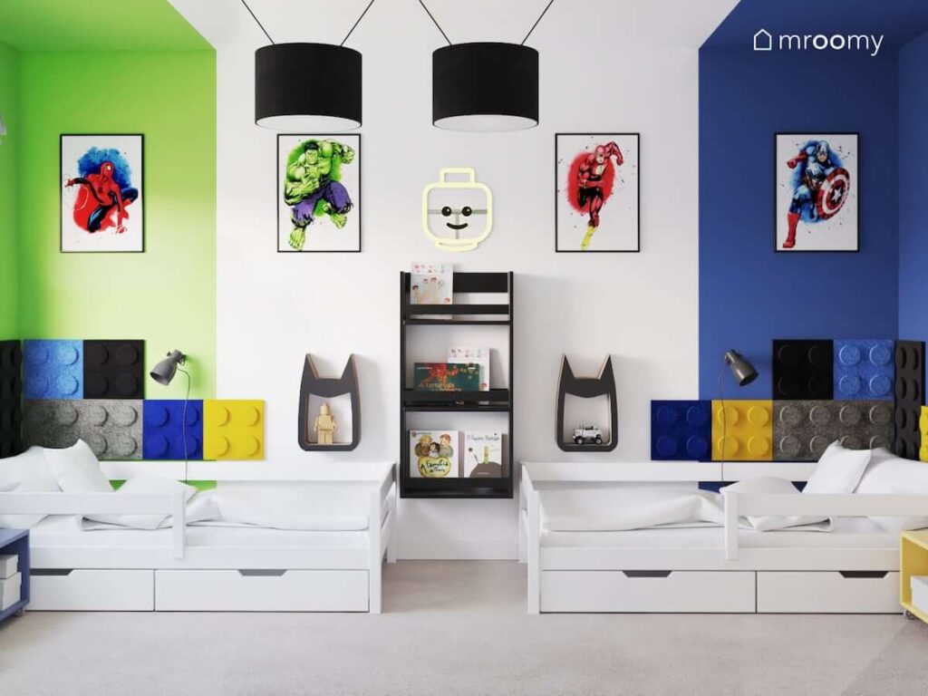 Dwa białe łóżka z szufladami oraz szafki ścienne w kształcie Batmana a także czarna biblioteczka i ozdobne plakaty z superbohaterami w pokoju dla dwóch chłopców