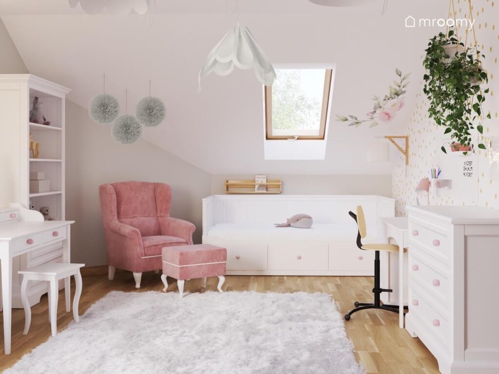 Jasny poddaszowy pokój dla dziewczynki z białymi meblami oraz różowym tapicerowanym fotelem oraz ozdobnymi pomponami lampami bezami i puszystym dywanem