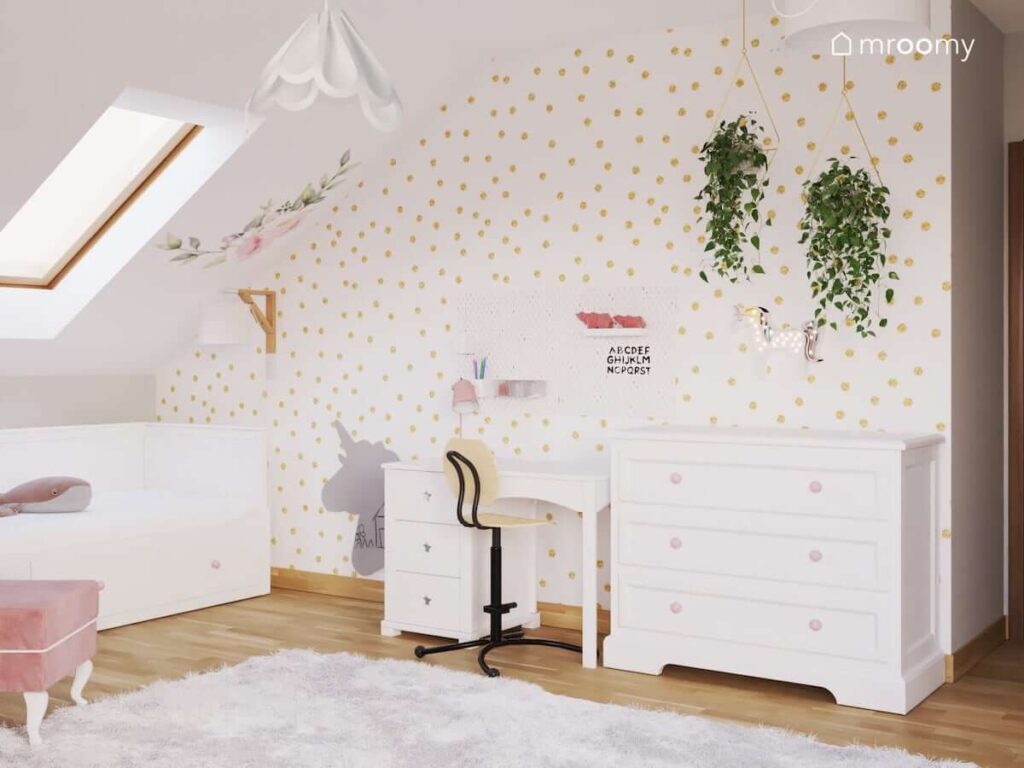 Poddaszowy pokój dla dziewczynki w wieku przedszkolnym z białym biurkiem organizerami ściennymi i komodą oraz tapetą w złote kropki i kwietnikami