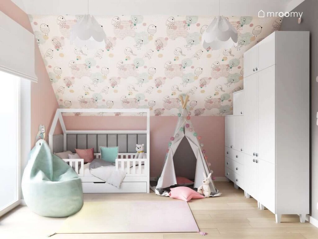 Pastelowo różowy pokój dla dziewczynki z łóżkiem domkiem uzupełnionym szarymi panelami ściennymi a także namiotem tipi i wygodną miętową pufą