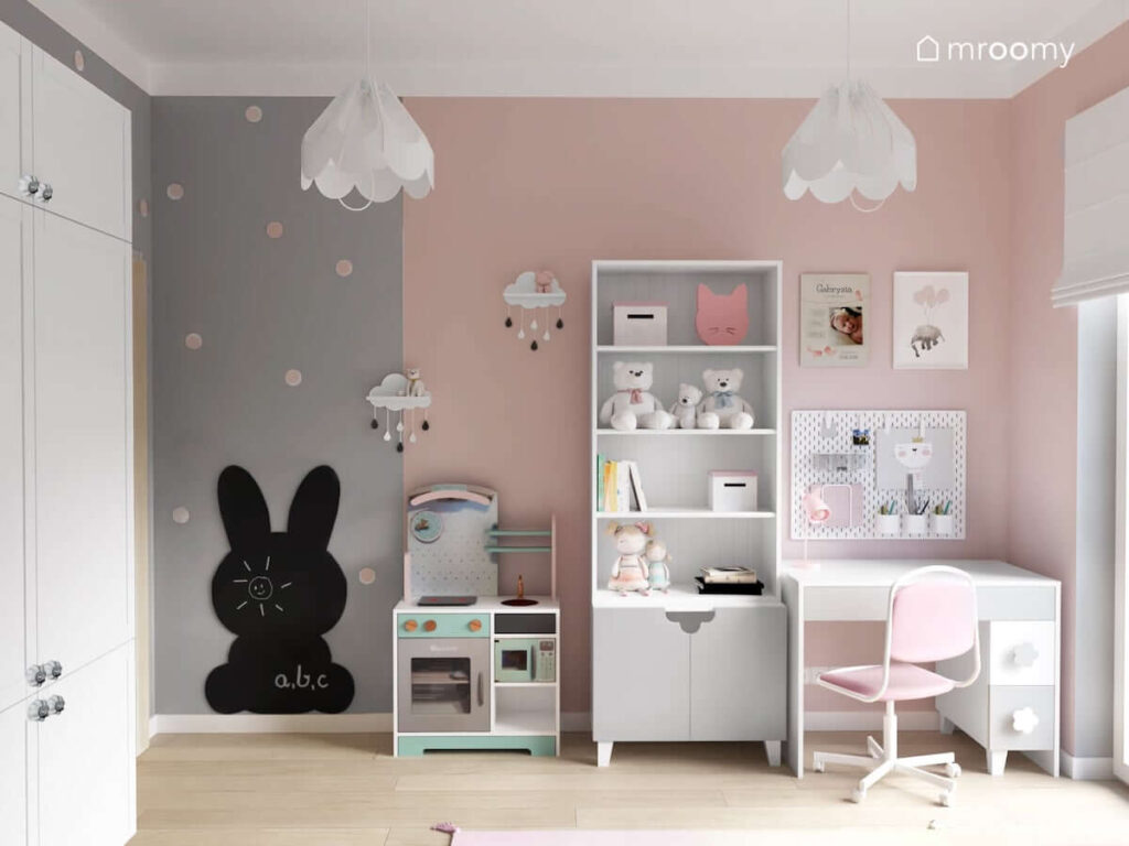 Białe biurko regał oraz kuchenka do zabawy a także tablica kredowa w kształcie królika oraz ozdobne lampy bezy w szaro różowo białym pokoju dla dziewczynki