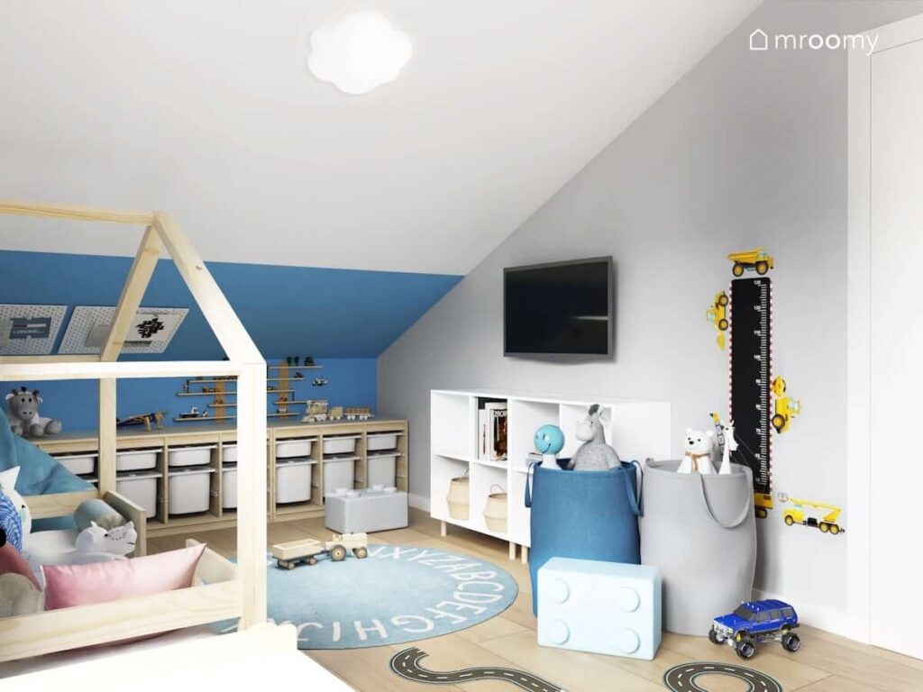 Niebieska część pomieszczenia w pokoju dla chłopca i dziewczynki z drewnianymi regałami z pojemnikami koszami na zabawki oraz pufkami w kształcie klocków i miarką wzrostu z motywami budowy