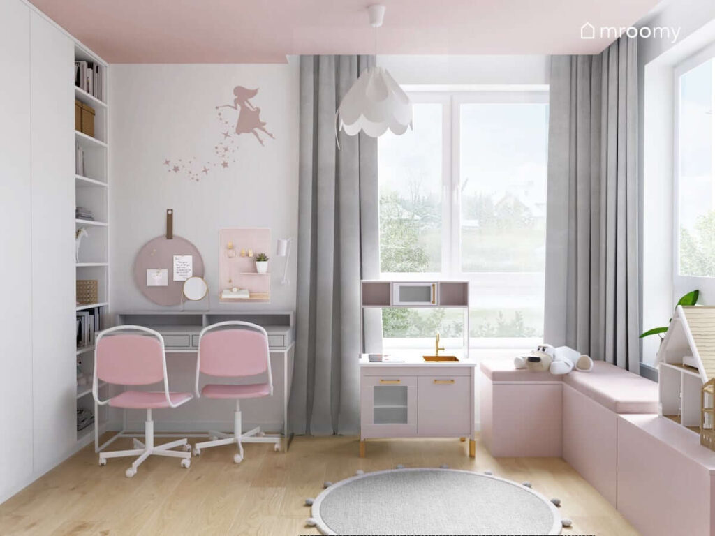 Biało różowy pokój zabaw dla dwóch sióstr z biurkiem z nadstawką i organizerami a także z kuchenką dla dzieci i różowymi skrzyniami na zabawki