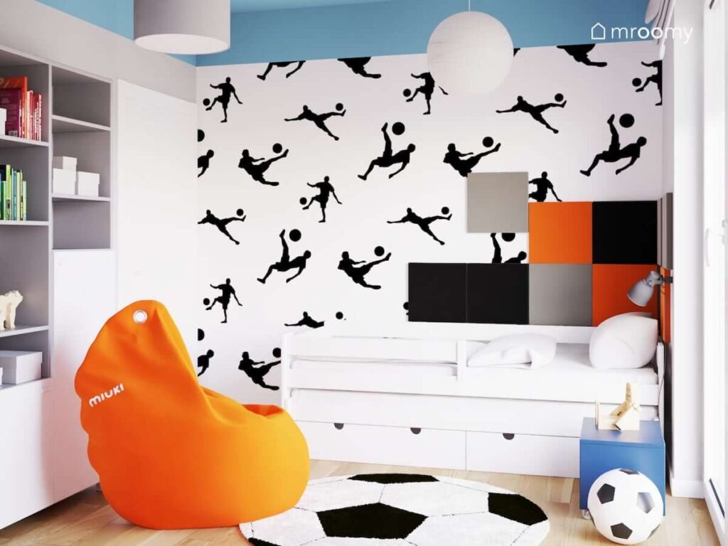 Białe łóżko w pokoju dla kilkuletniego chłopca a za nim tapeta z piłkarzami oraz czarne szare i pomarańczowe panele ścienne