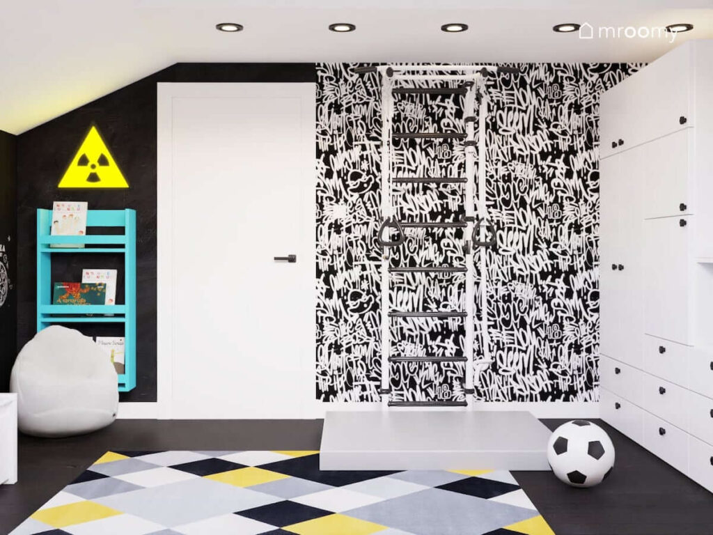 Ściana ozdobiona tapetą w graffiti a na niej drabinka gimnastyczna z materacem w biało czarnym pokoju dla chłopca