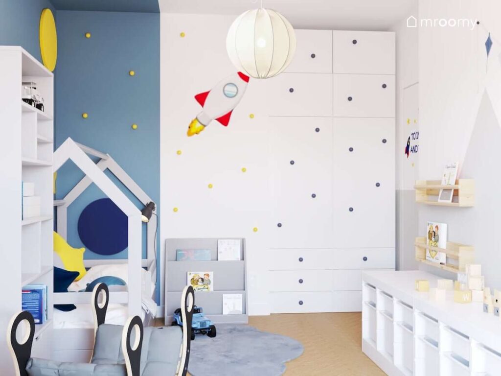Kosmiczny pokój dla malutkiego chłopca z dużą białą szafą uzupełnioną niebieskimi gałkami do mebli łóżkiem w kształcie domku ścianą uzupełnioną żółtymi gałkami oraz z naklejką w kształcie rakiety