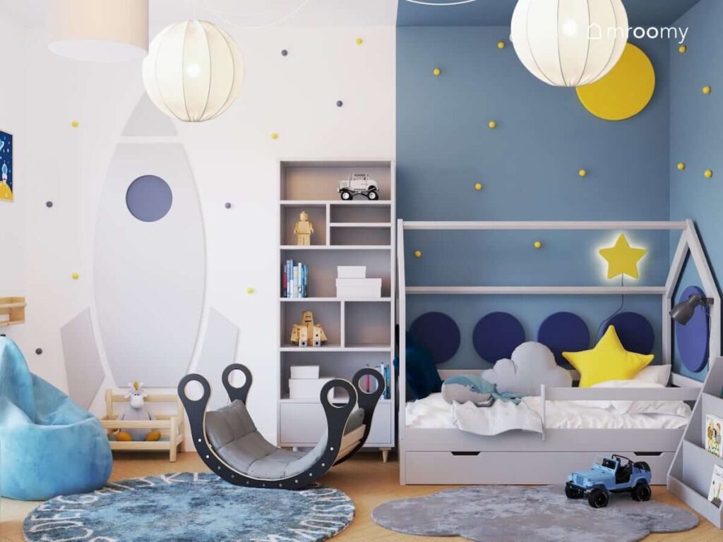 Biało niebieski pokój dla niemowlaka z czarnym bujakiem tablicą kredową w kształcie rakiety oraz z łóżkiem w kształcie domku uzupełnionym panelami ściennymi oraz gwiezdnymi dodatkami